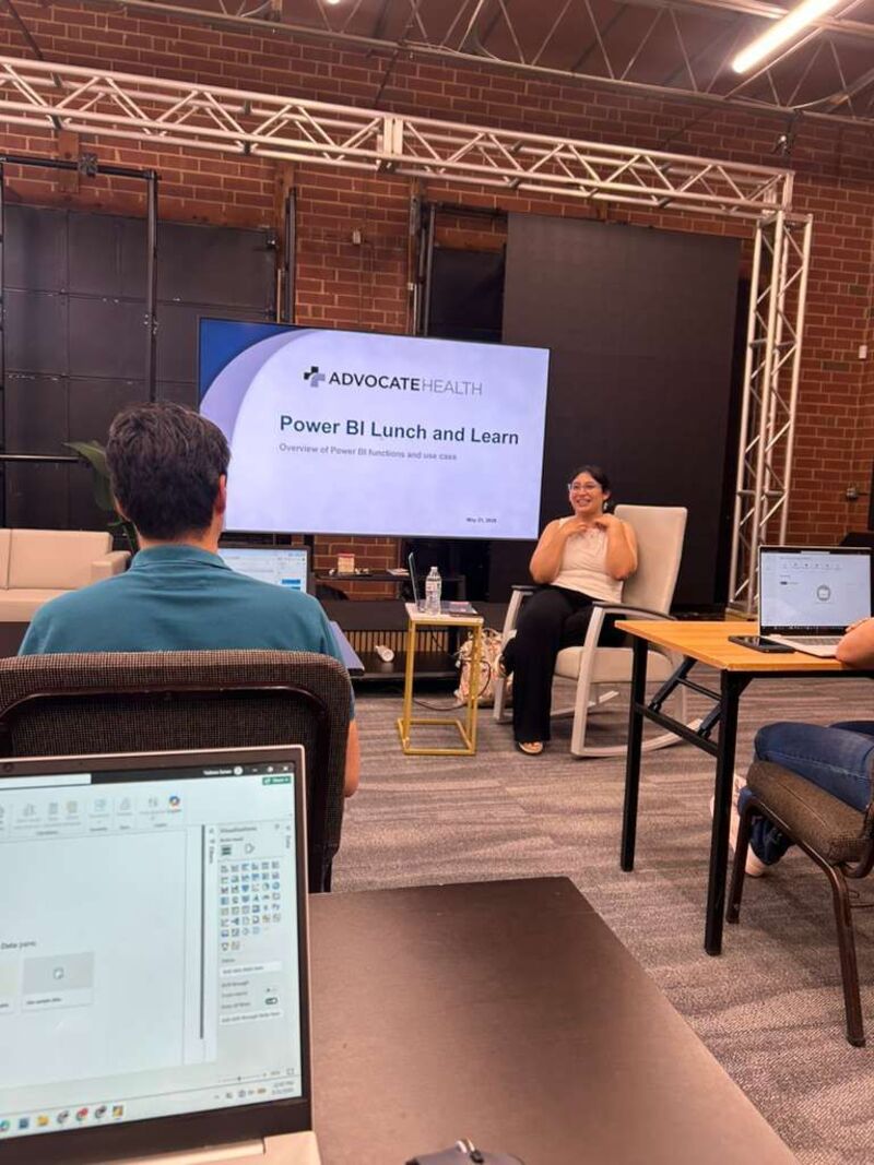 Sarai Ordonez smiling in a chair beside a screen reading Power BI Lunch and Learn, with attendees on laptops in the foreground